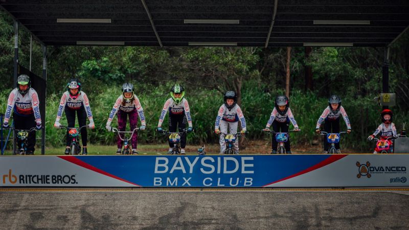 Eight junior riders lined up on their bikes with sign for club in front of them