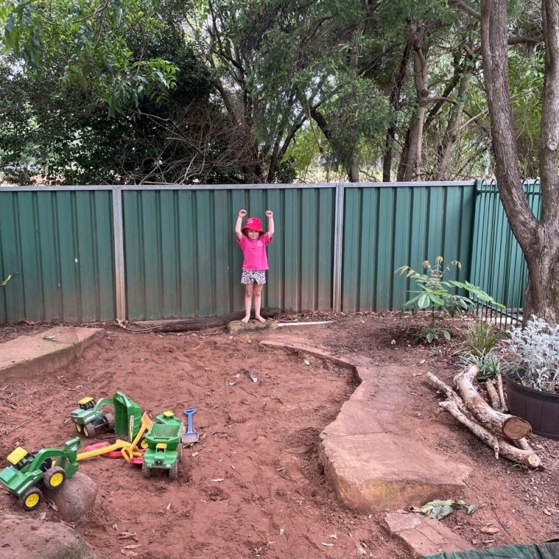 Child with arms up leaning against a fence at the back of a dirt garden